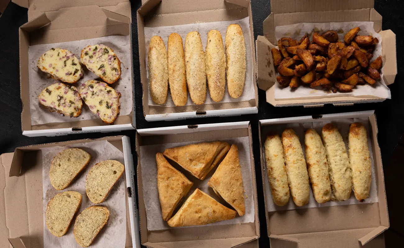 Assorted vegetarian sides in pizza boxes, including cheesy garlic bread, plain garlic bread, potato wedges, samosas, and stuffed breadsticks on a dark background