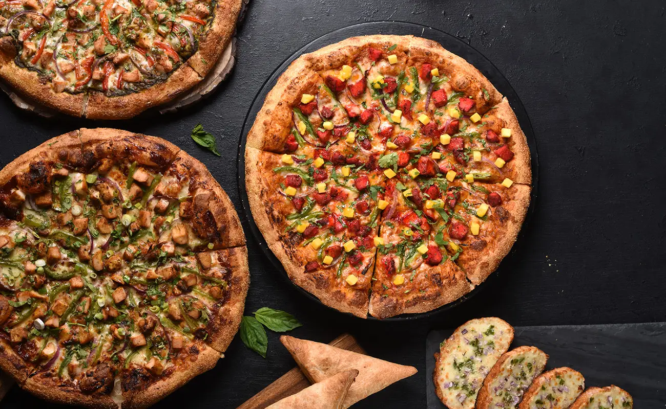 Three assorted vegetarian pizzas topped with colorful veggies, served with garlic bread slices and samosas on a dark background