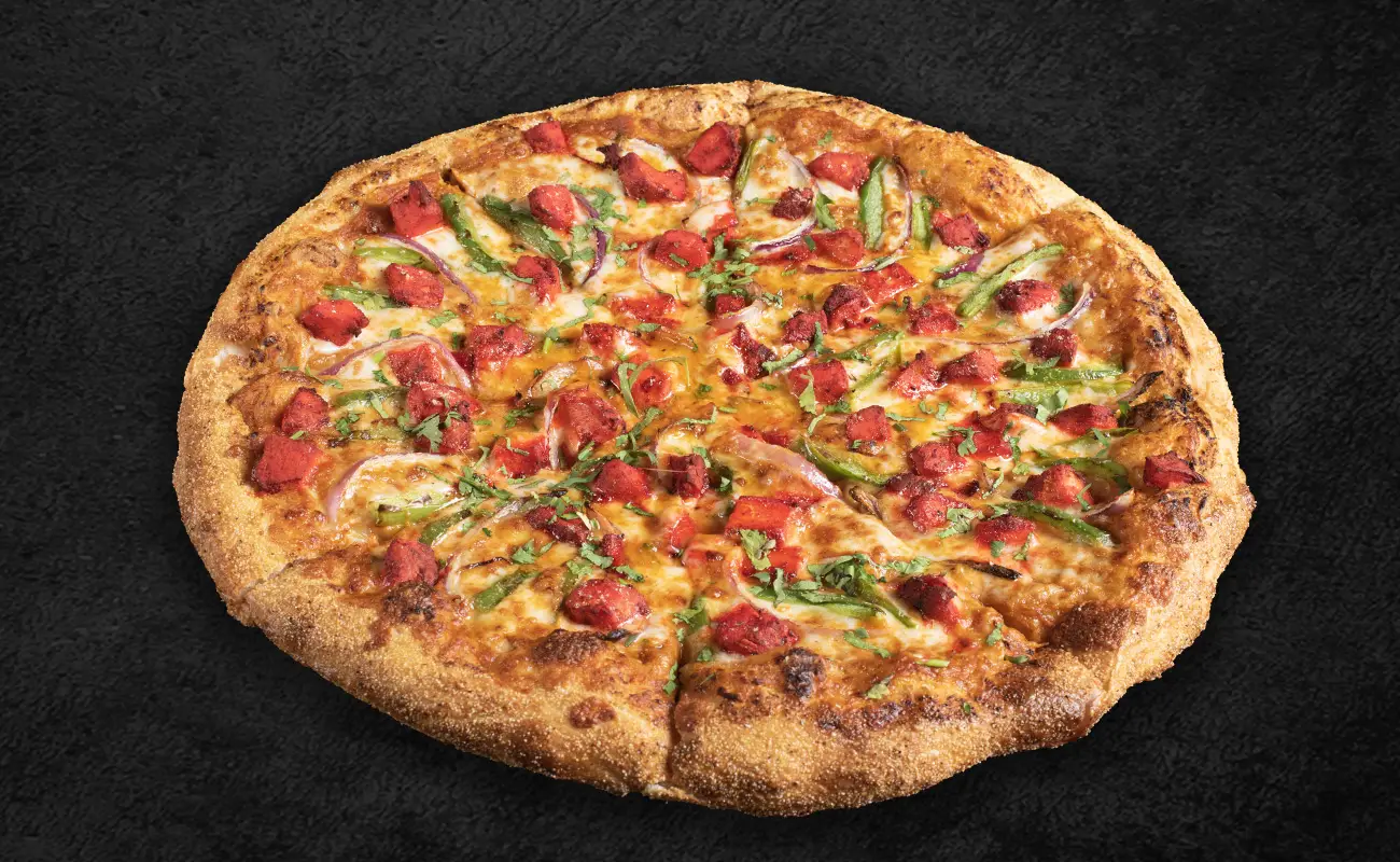 Tandoori-style pizza topped with marinated paneer chunks, green bell peppers, red onions, and fresh cilantro on a golden-brown crust, set against a dark background.