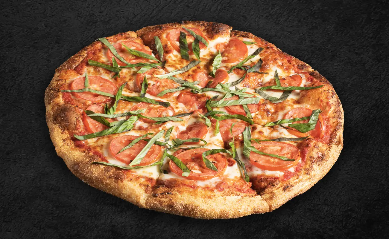 Fresh Margherita pizza topped with sliced tomatoes, shredded basil, and melted mozzarella cheese on a golden-brown crust, placed on a dark textured background