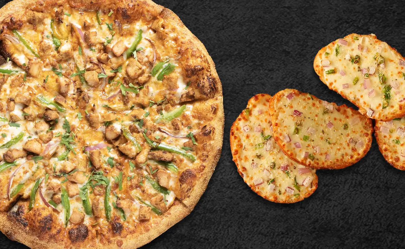 A close-up of a cheesy pizza topped with grilled chicken, green bell peppers, onions, and cilantro, placed next to several slices of garlic bread garnished with chopped onions and herbs on a dark background.