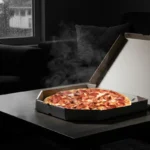 A hot, freshly delivered pizza in an open box sits on a coffee table in a dimly lit living room, with steam rising and a rainy window in the background.