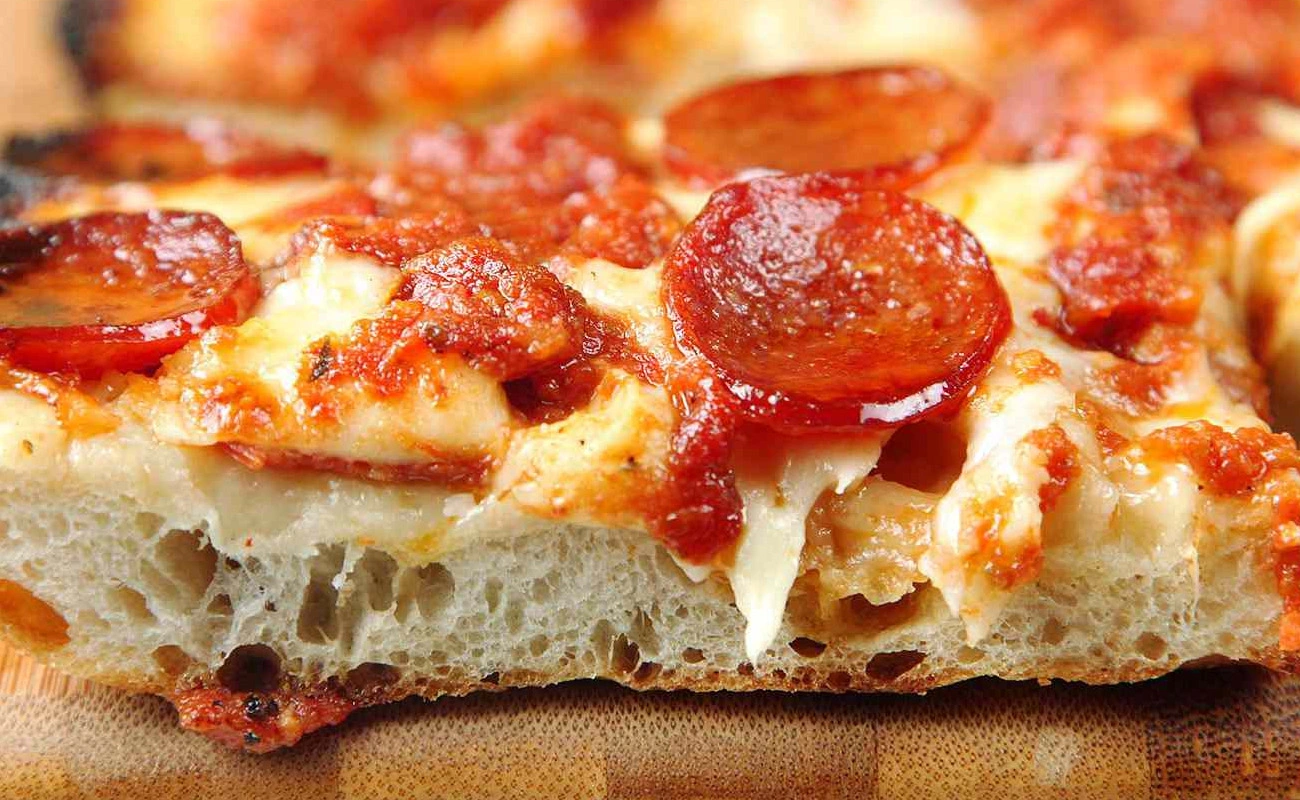 Close-up of Detroit-style pizza slice with crispy crust, melted cheese, tangy tomato sauce, and pepperoni toppings.
