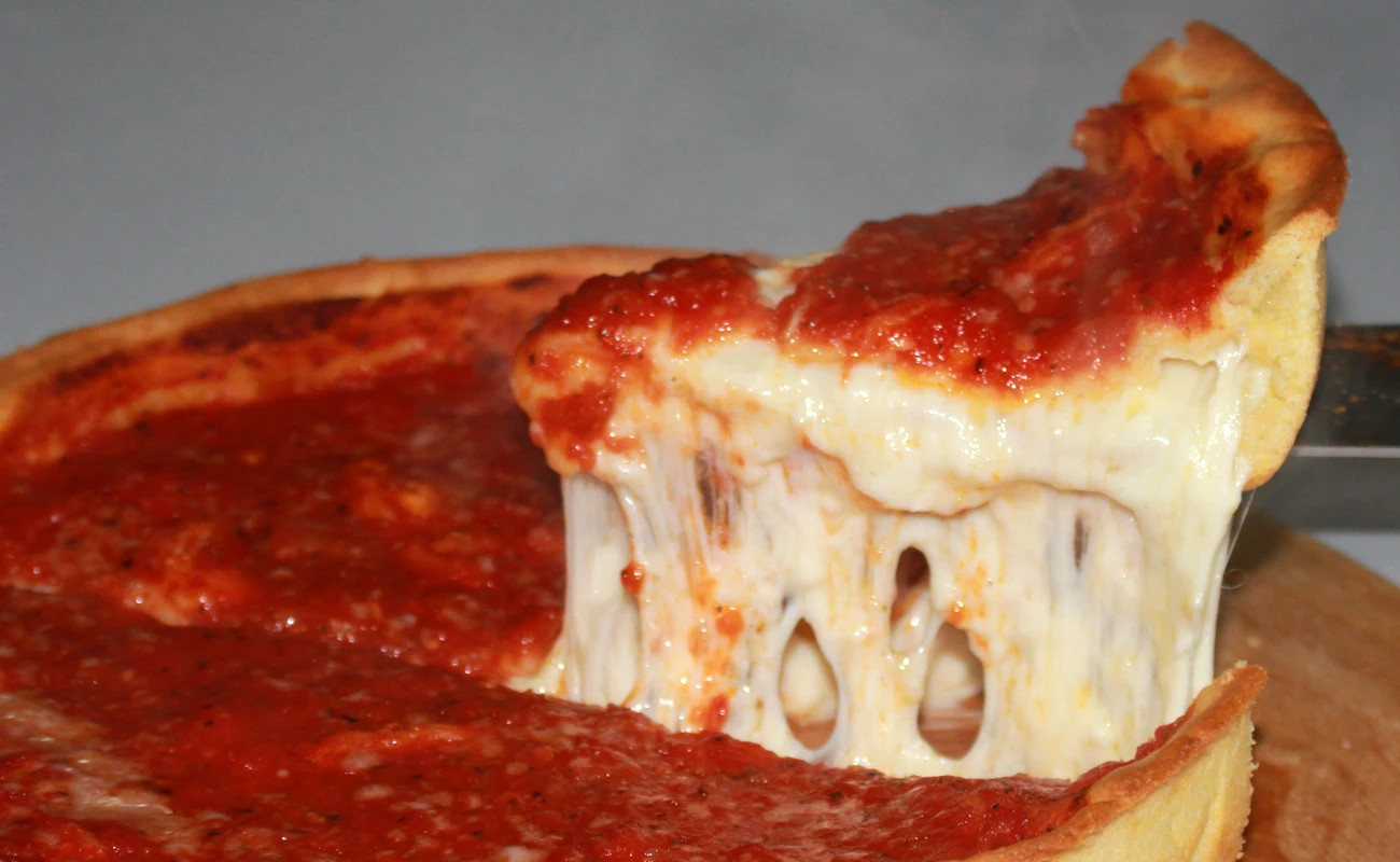 Close-up of a gooey Chicago-style deep-dish pizza with a thick crust, melted cheese, and rich tomato sauce, featuring a slice being pulled away on a wooden board.