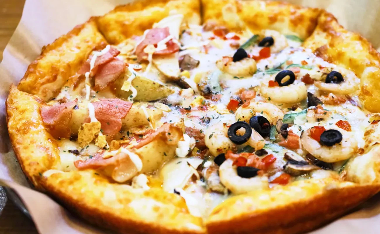 Colorful gourmet pizza with toppings like ham, olives, mushrooms, bell peppers, and creamy sauce on a thick crust, served on parchment paper for a delicious meal.