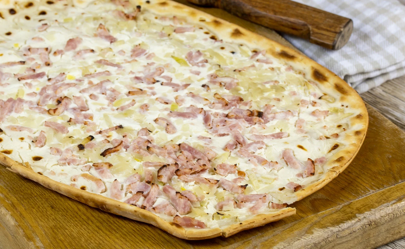 Traditional French flammkuchen with a thin crust topped with creamy sauce, caramelized onions, and crispy bacon, served on a wooden board with a checkered cloth.