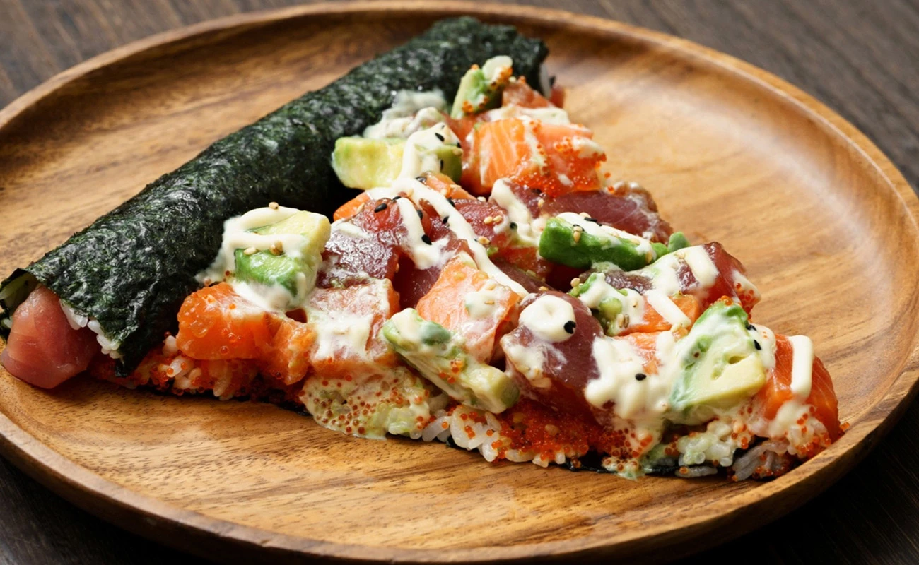 Unique sushi pizza slice with fresh salmon, tuna, avocado, and creamy sauce on a rice base, served alongside a seaweed-wrapped sushi roll on a wooden plate.