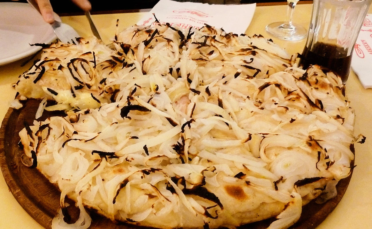 Traditional Italian focaccia bread topped with caramelized onions and melted cheese, served on a wooden board in a cozy restaurant with a glass of wine nearby.